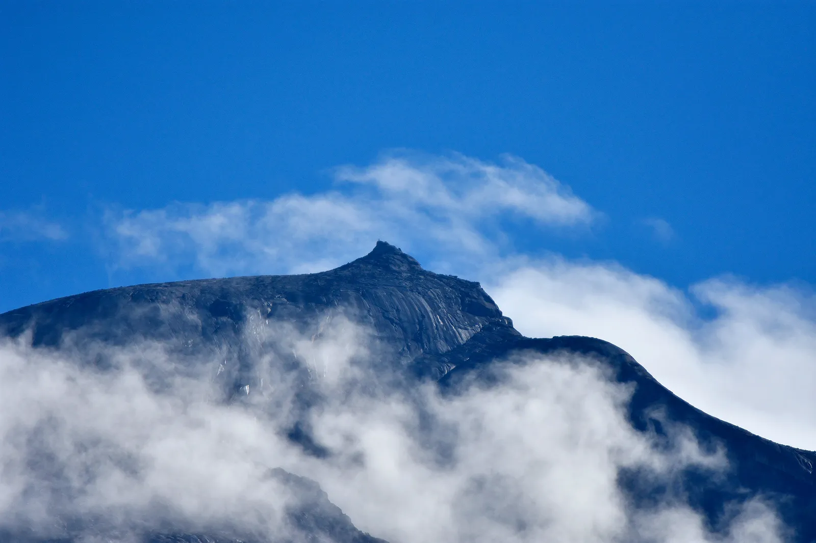 Summit of Mount Kinabalu rising out of cloud against a deep blue sky